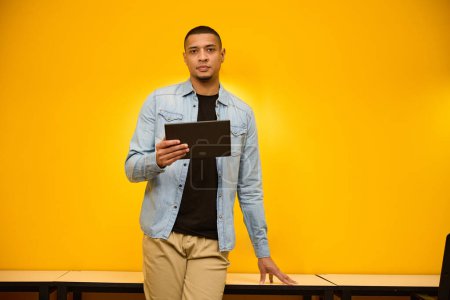 Téléchargez les photos : Copier la photo de l'espace de calme jeune homme afro-américain debout près du mur jaune dans le bureau tout en tenant la tablette dans la main - en image libre de droit