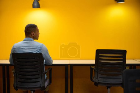 Photo for Back view picture of african american man sitting at table in office while turning his head to right - Royalty Free Image