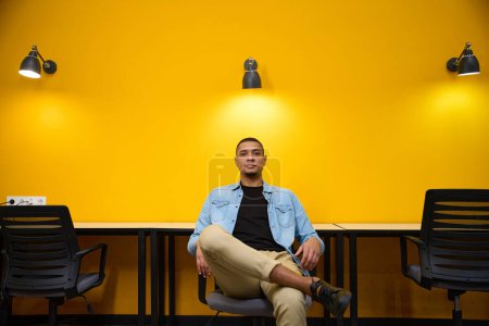 Téléchargez les photos : Calme jeune homme afro-américain gestionnaire assis sur la chaise près du mur jaune dans le centre d'appel tout en croisant ses jambes - en image libre de droit