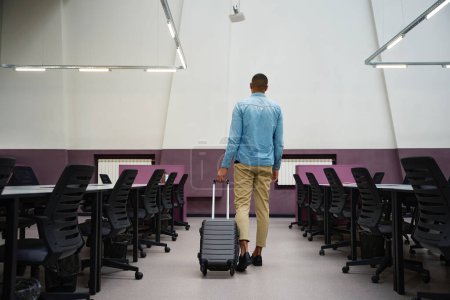 Photo for Back view photo of african american man standing in empty call center while holding suitcase by handle - Royalty Free Image