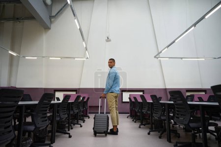 Photo for Calm young male employee standing between two rows of tables in empty coworking zone while holding suitcase in hand - Royalty Free Image