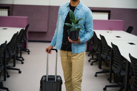 Photo for Cropped picture of man standing in coworking space while holding plant in flowerpot and rolling suitcase in hands - Royalty Free Image