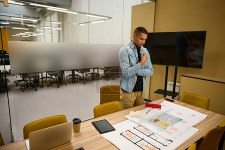Photo for Copy space photo of pensive young guy standing in office and looking at building plan which lying on table - Royalty Free Image