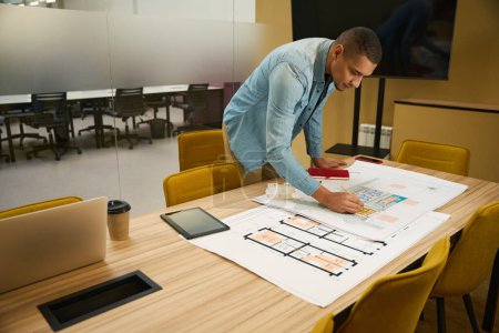 Foto de Arquitecto masculino joven enfocado apoyado sobre la mesa en el espacio de coworking mientras dibuja algo con lápiz en el plan de construcción - Imagen libre de derechos