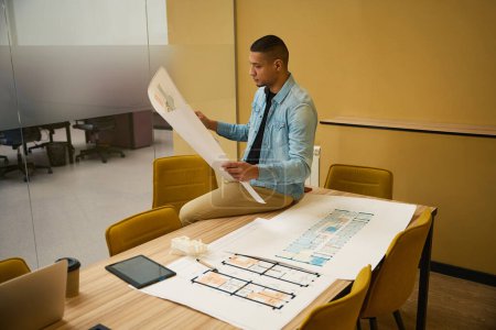Photo for Concentrated young man sitting on top of table in office while holding building blueprint in hands and looking at it - Royalty Free Image