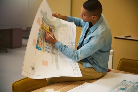 Photo for Serious young male architectural manager sitting on top of table and writing something with pencil on construction plan - Royalty Free Image