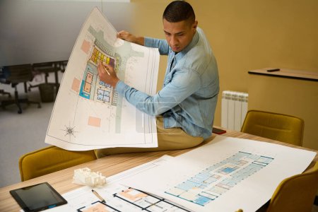 Photo for Serious young male employee correcting something on building plan which he holding in hand while sitting on top of table - Royalty Free Image