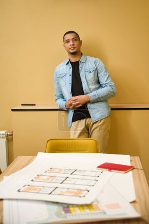 Photo for Calm young african american architectural manager standing in office near working place while leaning on wall ledge - Royalty Free Image