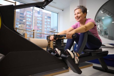 Photo for Adult smiling caucasian woman wearing sportswear doing sport exercise for back on rowing trainer in rehabilitation gym after injury. Concept of modern healthy lifestyle - Royalty Free Image