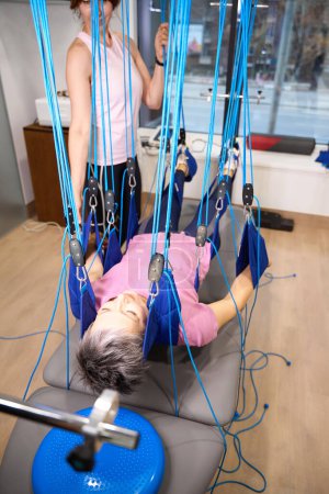 Photo for Obscure face female trainer helping middle aged woman hanging on suspension medical ropes after injury during recovery in rehabilitation center. Concept of modern healthy lifestyle - Royalty Free Image