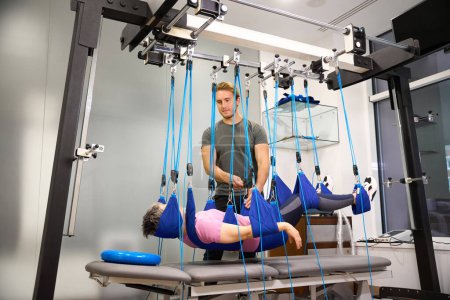 Photo for Young european male trainer helping middle aged woman hanging on suspension medical ropes after injury during recovery in rehabilitation center. Concept of modern healthy lifestyle - Royalty Free Image
