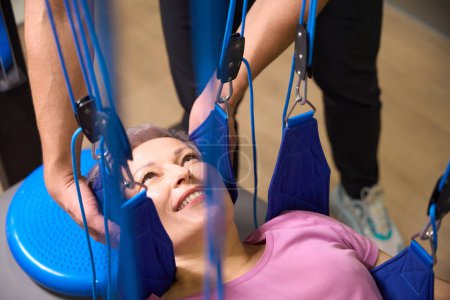 Photo for Cropped male trainer helping smiling mature caucasian woman hanging on suspension medical ropes after injury during recovery in rehabilitation center. Concept of modern healthy lifestyle - Royalty Free Image