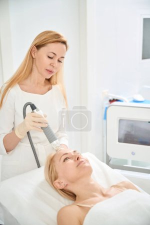 Photo for Skilled woman dermatocosmetologist conducting microneedle and radio frequency therapy, applying RF micro needle machine on clients forehead, aesthetic medicine clinic, lifting - Royalty Free Image