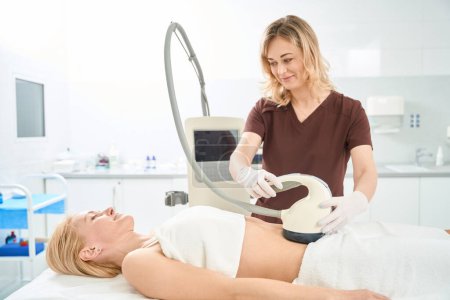 Smiling doctor conducting body contouring and cellulite reduction at abdomen zone to young woman patient, breaking down fat, stimulating collagen synthesis, improving skin texture