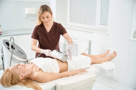 Femme cliente allongée sur le canapé médical alors qu'une dermatocosmétologue hautement qualifiée effectue une thérapie de rajeunissement qui lisse, tonifie, améliore la microcirculation et les formes