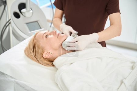 Infirmière professionnelle de clinique de médecine esthétique appliquant une thérapie endosphérique sur le visage du client féminin, stimulant la production de protéines d'élastine de jeunesse et de collagène dans la peau