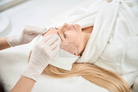 Photo for Skilled cosmetologist cleaning female client face from make-up and applying pore-expanding complex and moisturizer to dry and itchy skin, aesthetic medicine clinic - Royalty Free Image