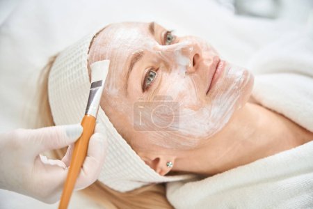 Cosmetologist in protective gloves applying collagen face mask to adult woman client face to hydrate and smooth facial wrinkles, accelerate the healing process of cracks and wounds