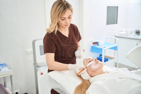 Skilled dermatocosmetologist keeping out care procedures on clients face, applying mask to improve skin condition, restore moisture balance and increase firmness and elasticity