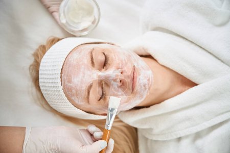 Top view leading expert in cosmetology applying alginate mask on female client face with help of silicone brush to improve skin condition, restore moisture balance and increase firmness and elasticity
