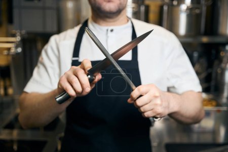 Téléchargez les photos : Couteau d'affûtage de chef masculin recadré avec aiguiseur sur cuisine floue au restaurant. Concept de saine alimentation savoureuse - en image libre de droit