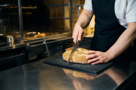Photo for Partial male chef cutting bread with knife on chopping board at table on blurred kitchen in restaurant. Concept of tasty healthy eating - Royalty Free Image