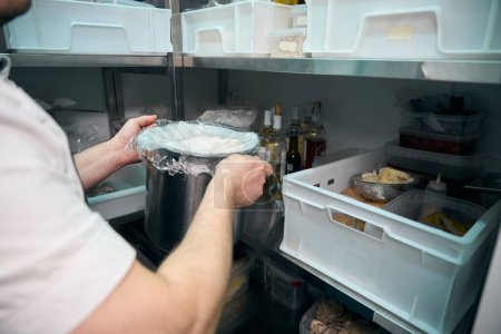 Téléchargez les photos : Chef masculin partiel mettre le plat dans une casserole sur l'étagère dans le réfrigérateur stockage dans le restaurant. Concept de saine alimentation savoureuse - en image libre de droit