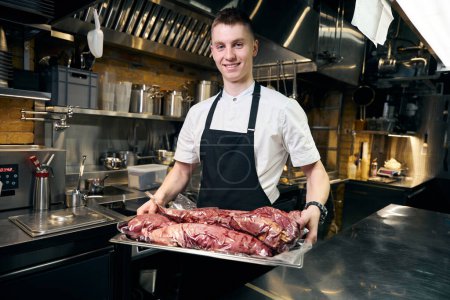 Photo for Smiling caucasian male chef holding iron tray with vacuuming fish and looking at camera on kitchen in restaurant. Concept of delicious healthy eating - Royalty Free Image