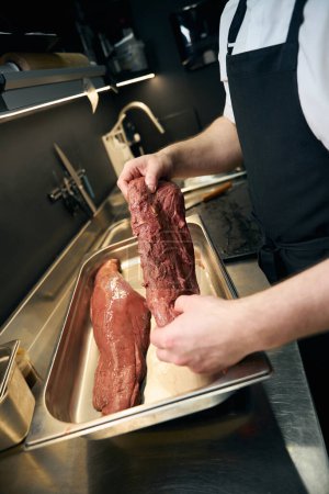 Téléchargez les photos : Chef masculin cultivé préparant filet de viande pour la cuisson sur un plateau de fer à table sur la cuisine dans le restaurant. Concept de saine alimentation savoureuse - en image libre de droit