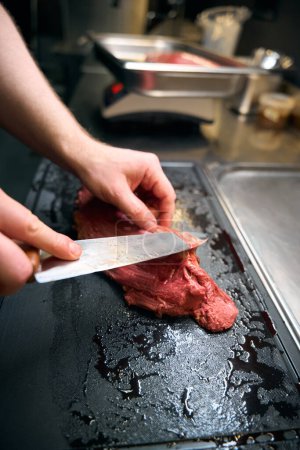 Téléchargez les photos : Chef mâle coupé filet de viande avec couteau sur planche à découper à table sur la cuisine dans le restaurant. Concept de saine alimentation savoureuse - en image libre de droit