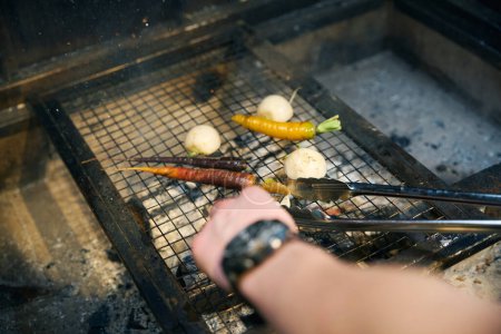 Cropped male chef smearing onion with brush and frying vegetables on iron net in burning fire place in restaurant. Concept of delicious healthy eating