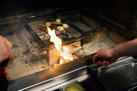 Partial male chef frying fish fillet on iron sticks and vegetables on iron net in burning fire place in restaurant. Concept of tasty healthy eating