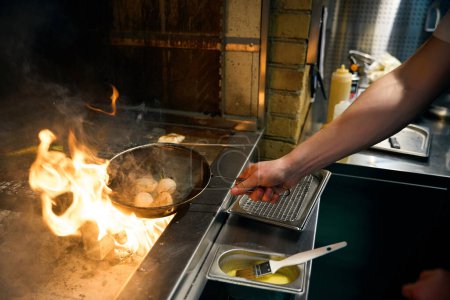 Side view of cropped male chef frying onions on kitchen grid and carrots on iron net in burning fire place in restaurant. Concept of delicious healthy eating