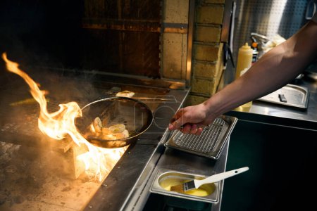 Side view of partial male chef frying onions on kitchen grid and carrots on iron net in burning fire place in restaurant. Concept of tasty healthy eating