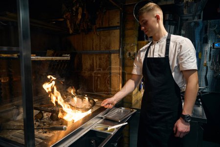 Téléchargez les photos : Chef caucasien faisant frire les oignons sur la grille de la cuisine et les carottes sur le filet de fer dans la cheminée en feu dans le restaurant. Concept de délicieuse alimentation saine - en image libre de droit
