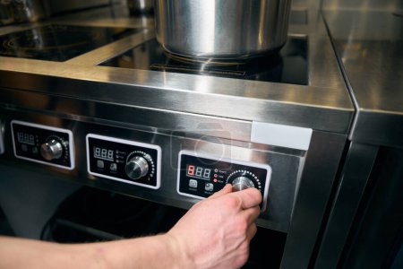 Téléchargez les photos : Coupé chef masculin allumant le bouton de la cuisinière électrique moderne pour chauffer le plat dans la casserole sur la cuisine dans le restaurant. Concept de saine alimentation savoureuse - en image libre de droit