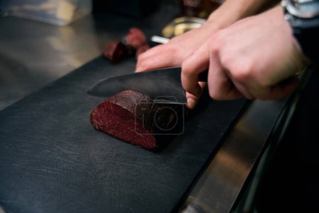 Téléchargez les photos : Concentration sélective du chef masculin cultivé coupant la viande avec un couteau sur la planche à découper à table sur la cuisine dans le restaurant. Concept de délicieuse alimentation saine - en image libre de droit