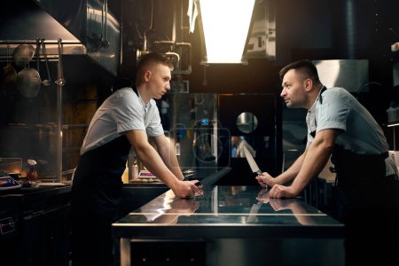 Photo for Side view of two serious caucasian male competitor chefs with knives looking at each other at table on kitchen in restaurant. Concept of tasty healthy eating - Royalty Free Image