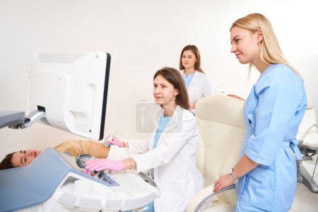 Photo for Female adult caucasian gynecologist doing ultrasound scanning of pregnant belly of young european woman while doctors colleagues looking at it in clinic. Concept of pregnancy and maternity - Royalty Free Image