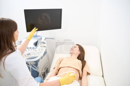 Téléchargez les photos : Gynécologue féminine faisant et montrant à la jeune femme européenne souriante ses résultats d'échographie du ventre enceinte dans une clinique moderne. Concept de grossesse et de maternité - en image libre de droit