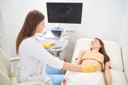 Photo for Female caucasian gynecologist doing ultrasound scanning of pregnant belly of young european woman and they looking at each other in modern clinic. Concept of pregnancy and maternity - Royalty Free Image