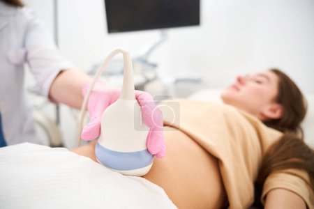 Téléchargez les photos : Gros plan de l'échographie du ventre d'une femme enceinte faisant gynécologue dans une clinique floue. Concept de grossesse et de maternité - en image libre de droit