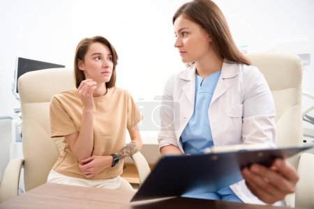 Foto de Mujer caucásica médico mostrando a la joven mujer europea preocupada sus resultados de examen y se miran entre sí en la clínica. Concepto de asistencia sanitaria - Imagen libre de derechos