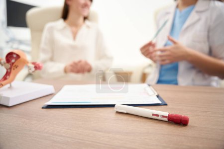 Focus on foreground of table with test, uterus model and clipboard with notes with blurred background of female gynecologist and woman patient in clinic. Concept of pregnancy and maternity