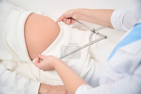Téléchargez les photos : Image recadrée d'une gynécologue féminine mesurant le ventre d'une femme enceinte avec étrier en clinique. Concept de grossesse et de maternité - en image libre de droit