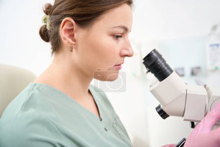 Foto de Vista lateral recortada de la doctora caucásica usando uniforme médico mirando en el microscopio durante el trabajo en la clínica. Concepto de asistencia sanitaria - Imagen libre de derechos
