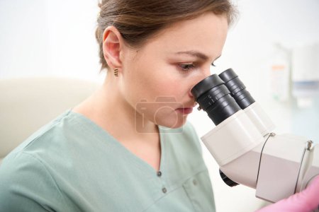 Foto de Vista parcial de la doctora caucásica enfocada usando uniforme médico mirando en el microscopio durante el trabajo en la clínica. Concepto de asistencia sanitaria - Imagen libre de derechos