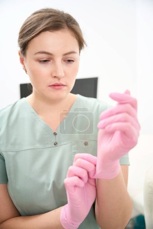 Téléchargez les photos : Une femme caucasienne met un gant médical à portée de main avant de travailler à la clinique. Concept de soins de santé - en image libre de droit