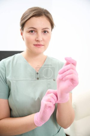 Téléchargez les photos : Médecin européen féminin mettant un gant médical sous la main avant le travail et regardant la caméra à la clinique. Concept de soins de santé - en image libre de droit