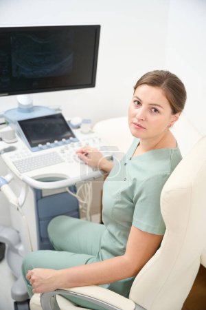Photo for Serious female caucasian doctor looking at camera while working on blurred modern ultrasound machine in clinic. Concept of healthcare - Royalty Free Image